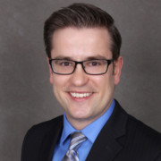 White man with brown hair and glasses smiling, wearing a suit with a blue shirt