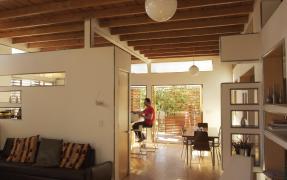 Inside of an eco-friendly home, showing a couch in the foreground, and a man sitting on a stool in the background