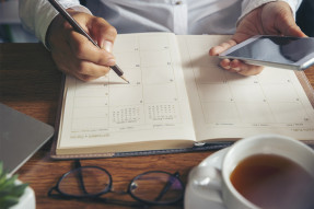 Person sitting at a desk holding a phone and writing in a planner