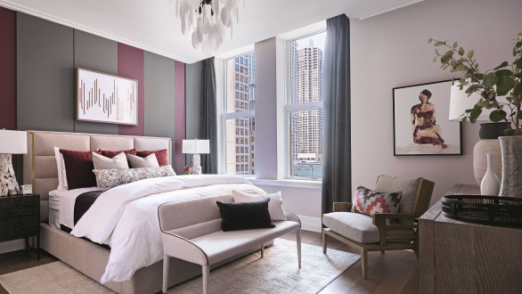 Photo of a bedroom with a queen sized bed, large window and chair. The bed is decorated with maroon, white, and beige linens. 