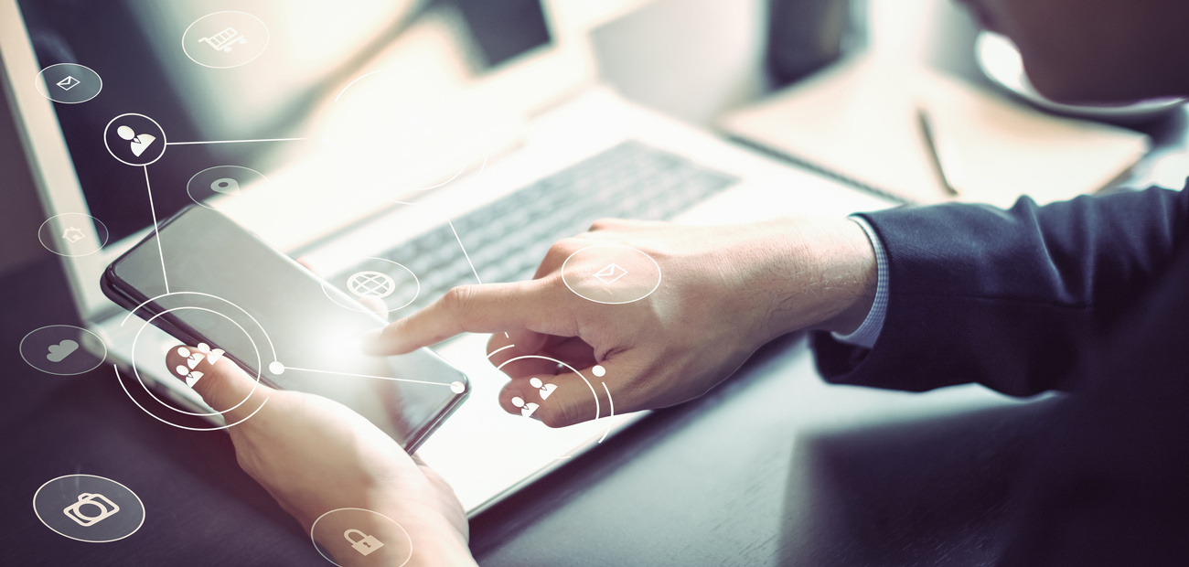 Man sitting at a desk and holding a cell phone with a laptop in the background