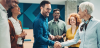 Two coworkers, a younger man and older woman, shake hands while smiling. A small group of coworkers stand with them