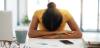 Brown woman with dark curly hair up in a bun with her head down on a desk