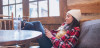 A young woman sits relaxed leaning back on a cushioned chair at a table while scrolling through her phone.