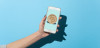 A person's arm and hand holding a smart phone displaying a bitcoin. The background is light blue with their shadow to the right.