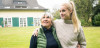 An elderly woman and a young adult woman stand with one arm around facing the camera looking up to their right in a large yard.