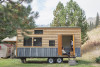 A picture of a tiny home on a wheeled trailer, parked on a grassy lot with hills, trees, and mountains in the background.