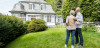 A retired couple, a man and a woman, stand on the right in a yard facing a new home in the background.