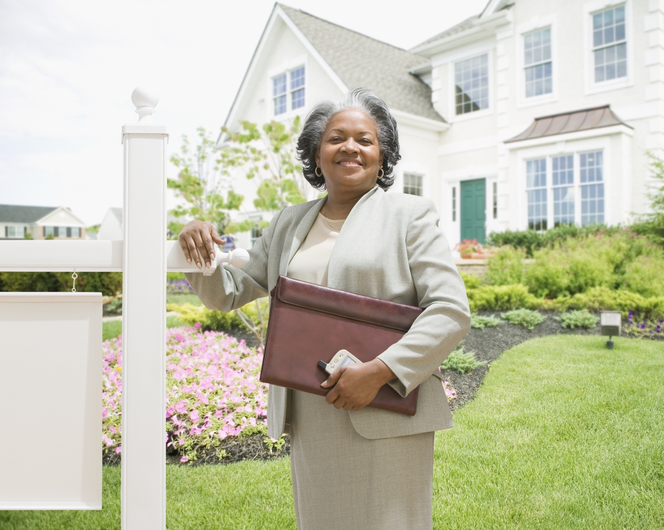 A woman standing in front of a house for sale sign, holding a briefcase and phone